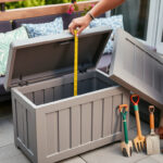 Cushions and garden tools beside a lidded outdoor storage box on a terrace, with a person checking measurements before buying.
